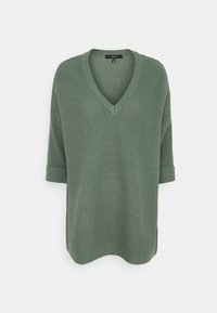 Maglione verde a V con maniche corte arrotolate, caratterizzato da un motivo tricot testurizzato simile a una waffle. Design leggero e casual.