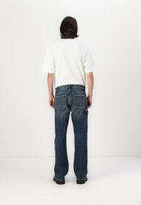 Jeans de ganga com corte reto, cor azul escura, dois bolsos traseiros e um leve efeito de desgaste; combinados com uma t-shirt branca simples.