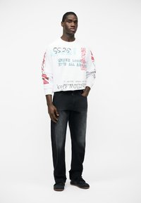 Homme portant un sweat-shirt blanc avec un motif et du texte, ainsi qu'un jean noir, debout avec une main dans la poche sur fond blanc.