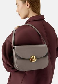 Borsa a tracolla in pelle grigia con chiusura a pattina e un disco di metallo dorato, indossata sulla spalla sopra una giacca bordeaux.
