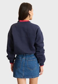 Marineblaues Sweatshirt mit lockerem Schnitt, rundem Ausschnitt und roten Rüschendetails am Kragen. Kombiniert mit einem kurzen Jeansrock mit Gesäßtaschen.