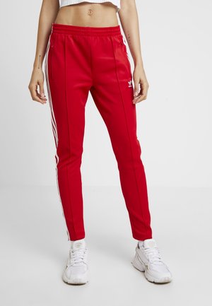 Femme portant un pantalon de survêtement rouge Adidas avec des bandes blanches et un logo, associé à des baskets blanches et un crop top blanc.