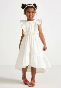 Vestito bianco in cotone con maniche arricciate, raccolto in vita e orlo a balze. Abbinato a sandali rossi e fascia floreale per capelli.