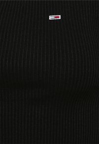 Svart ribbad topp med vertikal struktur. Litet broderat logotyp med röda, vita och blå accenter på vänster bröst.