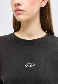 Γυναίκα με μακριά καστανά μαλλιά που φοράει μαύρο μπλουζάκι με στρογγυλή λαιμόκοψη και μικρό λευκό γεωμετρικό λογότυπο στο στήθος.