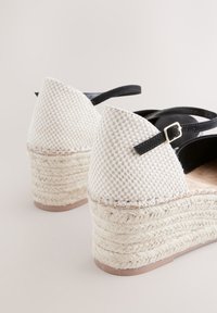 Keil-Espadrille-Sandalen mit geflochtenen Jutestöckeln, weißen gewebten Obermaterialien und schwarzen Knöchelriemen mit kleinen goldenen Schnallen.