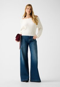 Pull crème en maille torsadée, à col bateau, associé à un jean denim bleu évasé. Accessoirisé avec une pochette bordeaux et des chaussures noires.