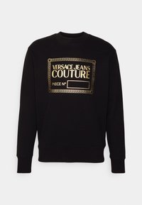 Svart sweatshirt av mjukt tyg. Har en guldfolie-logotryck "VERSACE JEANS COUTURE" i rektangulär design. Ribbad mudd.