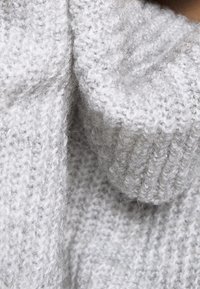Světle šedý pletený svetr s hrubou žebrovanou strukturou. Má vysoký límec a překládaný límec pro více tepla a pohodlí.
