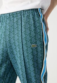 Pantalons de sport verts à motifs avec des rayures latérales bleues et blanches, arborant un logo crocodile Lacoste sur le côté gauche.