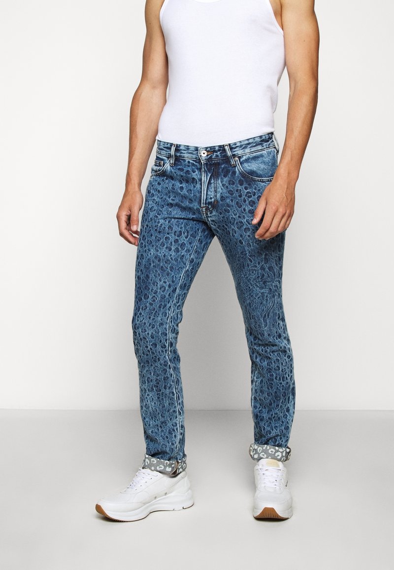 Niebieskie dżinsy z fakturowanym wzorem, zwężanym krojem, pięcioma kieszeniami oraz zawiniętymi mankietami z kontrastowym nadrukiem. Noszone z białymi sneakersami.
