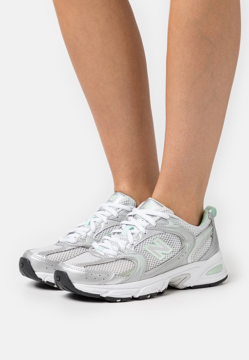 Zapatillas deportivas plateadas con material de malla, cordones blancos y detalles verdes. Cuentan con una suela acolchada y paneles laterales texturizados.