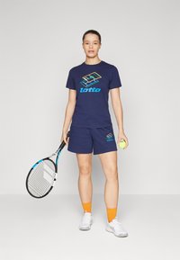 Mørkeblå Lotto trenings T-skjorte og shorts, logo i turkis og neonfarger, holder en gul tenniskule og racket, hvite sneakers, oransje sokker.
