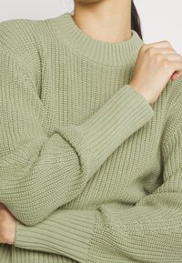 Pull tricoté vert clair avec un col rond, des poignets côtelés et un tissu texturé, présentant une coupe ample et une apparence confortable.