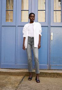 Grå denimjeans med rak passform, tillsammans med en vit stickad topp och en överdimensionerad vit skjorta. Svarta sandaler fulländar outfiten. Bakgrund med en blå dörr.