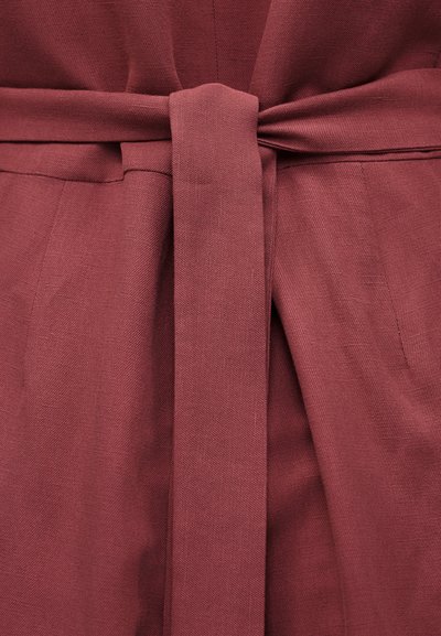 Κοντινό πλάνο ζωνής από ύφασμα που δένεται σε αδιάλειπτο κόμπο πάνω σε ρουχισμό με υφή και σε χρώμα σκόνης ρόδου.