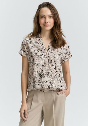 Camicia a maniche corte in tessuto beige chiaro, con un motivo a macchie nere, chiusura con bottoni e vestibilità rilassata. Abbinata a pantaloni chiari.