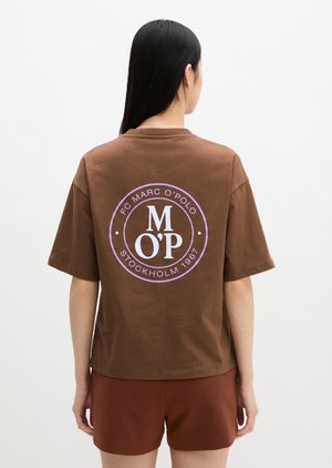 Femme aux longs cheveux noirs porte un t-shirt marron avec un logo circulaire violet et un short marron assorti, debout devant un fond blanc uni.