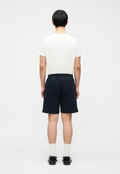 Άνδρας που στέκεται με την πλάτη προς τα εμάς, φοράει λευκό μπλουζάκι, μαύρο σορτς, λευκές κάλτσες και μαύρα παπούτσια, πάνω σε ένα απλό φωτεινό φόντο.