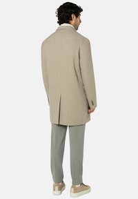 Beiger Wollmantel mit strukturierter Oberfläche, einfacher Rückenschlitz und spitze Reverskragen; kombiniert mit hellgrauen Hosen und cremefarbenen Sneakers.