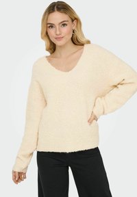 Cremefarbener, weicher Strickpullover mit V-Ausschnitt und überschnittenen Schultern, der eine lässige Passform und einen fransigen Saum aufweist. Kombiniert mit schwarzen Hosen.