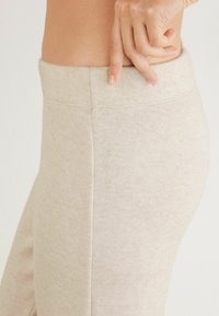 Beige legging met een textuurstof, hoge taille en zonder zichtbare naden, met een close-up van de tailleband en een hand die de pasvorm aanpast.