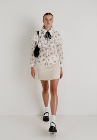 Blusa de chiffon floral branca com gola rendada, combinada com uma saia mini bege. Uma mala preta e sapatos com meias brancas completam o visual.
