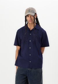 Camisa de manga corta azul marino con bordado floral en el pecho, cierre de botones en la parte delantera y un corte relajado, combinada con pantalones de mezclilla oscuros y una gorra beige.