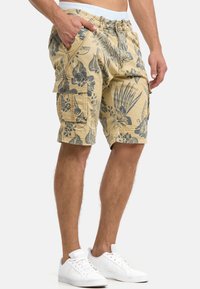Shorts cargo jaunes avec un motif floral bleu foncé, comportant des poches latérales et une taille à cordon. Portés avec des baskets blanches.