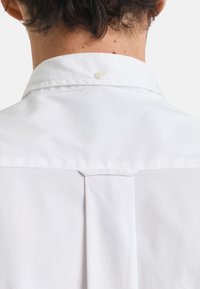 Weißes Hemd mit Knopfleiste, ausgestattet mit einem klassischen Kragen, einer Rückenfalte und einer subtilen Stoffstruktur. Details umfassen einen einzelnen Knopf an der Basis des Kragens.