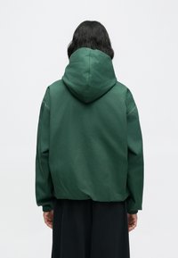 Veste à capuche verte à coupe décontractée, dotée de poignets élastiques et d'une texture lisse. Détail notable d'une couture au dos et design sobre.