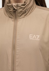 Μπεζ μπουφάν με φερμουάρ και ψηλό γιακά, με το λογότυπο EA7 Emporio Armani τυπωμένο στο αριστερό μέρος του στήθους.