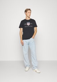 Svart bomull T-skjorte med "GANT"-logo og røde detaljer, sammen med lyseblå denimjeans og hvite joggesko. Enkel, uformell design.