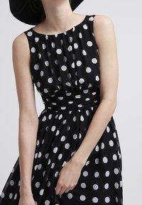 Robe sans manches noire avec des pois blancs, corsage froncé et taille cintrée. Tissu léger avec une texture lisse.