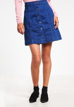 Kvinde iført en blå denimnederdel med knapper foran, pink langærmet top og sorte ankelstøvler, stående mod en hvid baggrund.