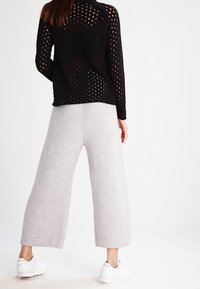 Svart öppet stickad tröja med långa ärmar, tillsammans med ljusgrå vida byxor. Vita sneakers syns, bakgrunden är enkel och ljus.