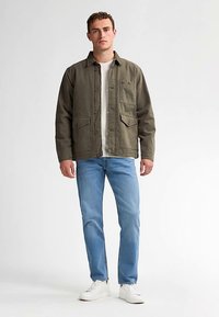 Giacca verde oliva con chiusura a bottoni, tasca sul petto e due tasche inferiori, abbinata a jeans azzurri e sneakers bianche.