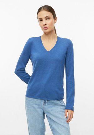 Vrouw met bruin haar in een paardenstaart, gekleed in een blauwe trui met lange mouwen en V-hals en lichtblauwe jeans tegen een witte achtergrond.