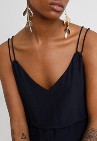 Granatowa sukienka z cienkimi ramiączkami i dekoltem w kształcie litery V. Złote kolczyki w kształcie liści mają teksturowaną powierzchnię z wieloma warstwami. Widoczna gładka tekstura skóry.