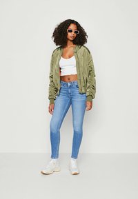 Olivgrön bomberjacka, vit kort topp, blå skinny jeans, vita sneakers med beige detaljer, stiliserat med runda solglasögon.