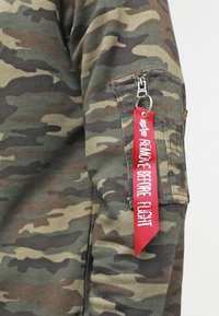Camisa de manga larga con patrón de camuflaje en tonos verdes y marrones, que presenta un bolsillo con cremallera y una etiqueta de tela roja que dice "RETIRAR ANTES DEL VUELO".