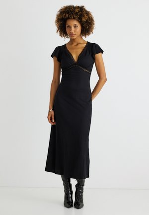 Frau mit lockigem Haar, die ein eng anliegendes schwarzes Midikleid mit Spitzenbesatz und schwarze Stiefel mit Absatz trägt, steht vor einem schlichten Hintergrund.