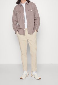 Camicia button-up marrone chiaro con due tasche sul petto, t-shirt bianca sotto, pantaloni beige e sneakers crema con dettagli azzurri.