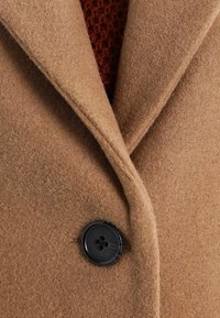 Cappotto di lana marrone con rever a notched, un bottone nero e una finitura morbida e strutturata, indossato sopra un indumento di maglia rosso scuro.