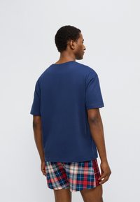 Μπλε βαθυά βαμβακερό κοντομάνικο μπλουζάκι με στρογγυλή λαιμόκοψη και κοντά μανίκια. Συνδυασμένο με καρό σορτς σε κόκκινο, άσπρο και μπλε. Το μοντέλο έχει γυρισμένη την πλάτη.