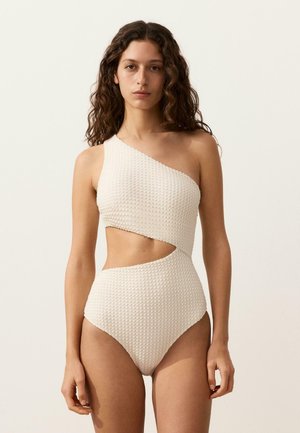 Žena s kučeravými vlasmi, ktorá má na sebe textúrované jednoramenné plavky s diagonálnym výrezom v oblasti pásu, stojaca na pozadí svetlej jednoliatej steny.