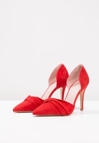 Une paire de chaussures à talons hauts en daim rouge avec des pointes et un design plissé à l'avant, posée sur une surface blanche contre un fond uni.