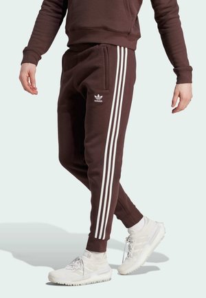 Osoba ubrana w brązowe spodnie dresowe Adidas z trzema białymi paskami i białe trampki, stojąca na jednolitym jasnym tle.