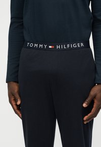Person iført sorte bukser med et synligt Tommy Hilfiger-logo i linningen og en mørk langærmet skjorte, hænderne hviler ved hofterne.