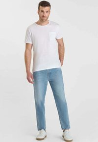 T-shirt bianca in cotone con taschino sul petto e jeans denim azzurri. Il modello indossa sneakers bianche con dettagli beige. Design minimalista.